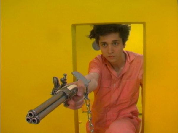 Film still of a person wearing a peach jumpsuit and one handcuff crouching in the small doorway of a mustard yellow room and aiming a three barrel gun to the left of the camera