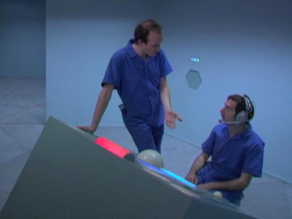 Film still of two men, one standing and one seated, dressed in blue jumpsuits talking near a futuristic console