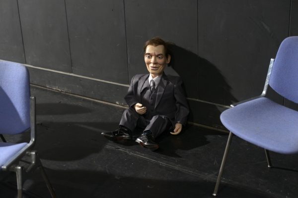 Ventriloquist puppet of a man dressed in suit sitting with its back against a black wall