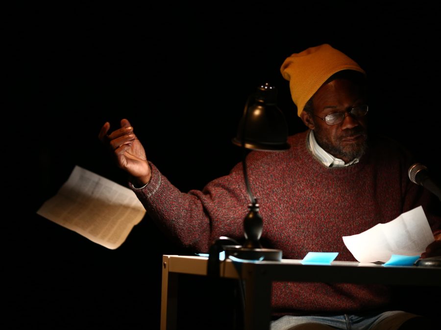 A dark-skinned older man sits at a table illuminated by a desk lamp on an otherwise pitch-black stage. He looks at one sheet of paper and tosses another to the floor.
