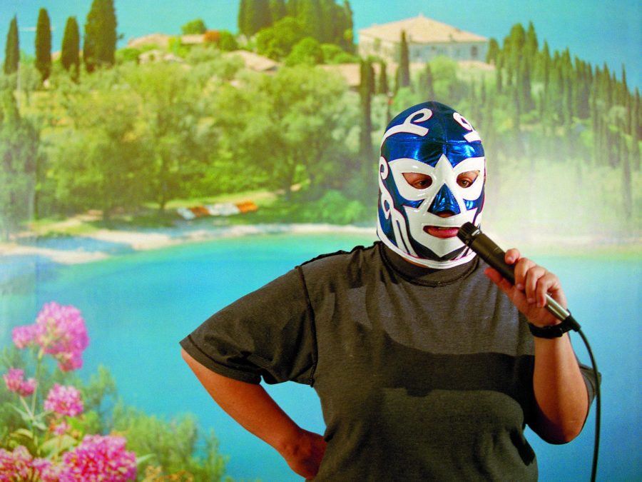 Against a tropical backdrop, a person in a blue-and-white wrestling mask holds a microphone up to their mouth.