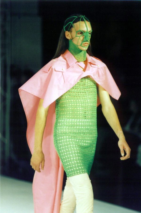 A runway model with a pink cape and a green crocheted dress that connects to a mask covering the neck and nose
