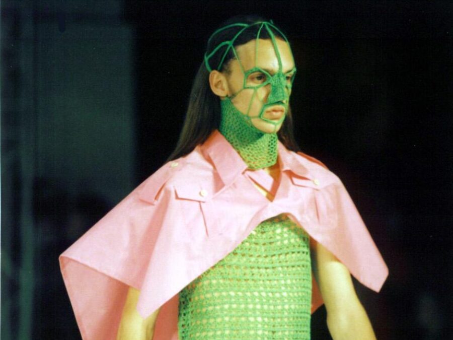 A runway model with a pink cape and a green crocheted dress that connects to a mask covering the neck and nose