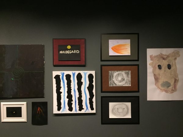 Several paintings and drawings of different sizes and in different colored matted frames on a dark gray wall