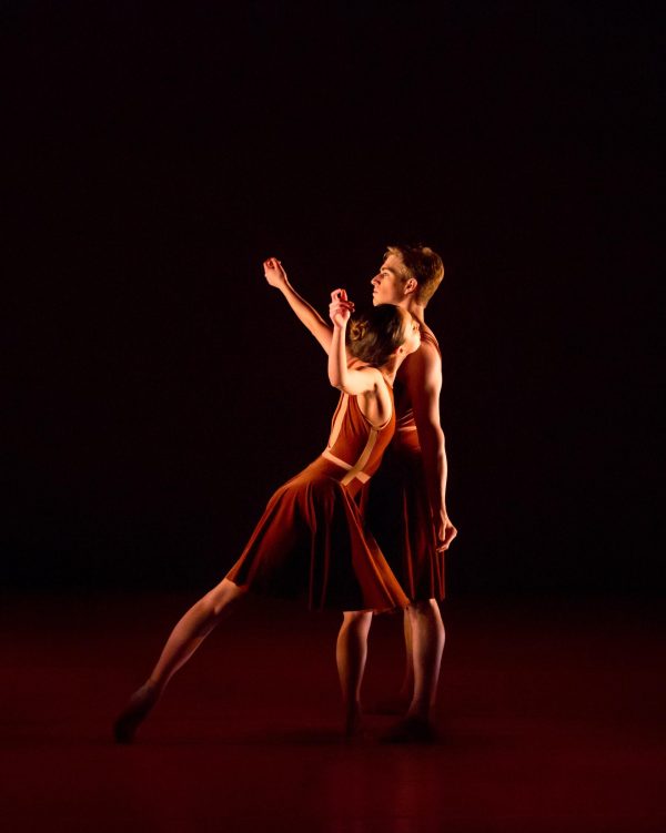 Two dancers lean on each other in a dimly lit theater space with a black backdrop