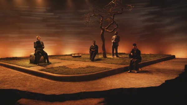 Four people dispersed among a wasteland setting backlit by orange lighting; three people sit and one stands next to a dead tree.
