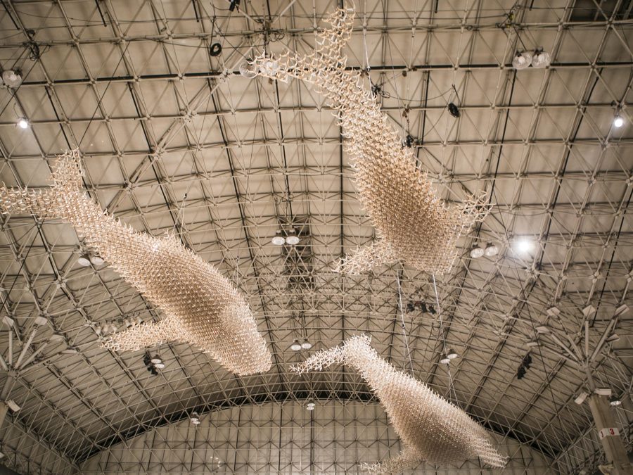 Three whale or giant fish forms, made of light tan colored sticks of some kind, are suspended from the ceiling of a large warehouse space.