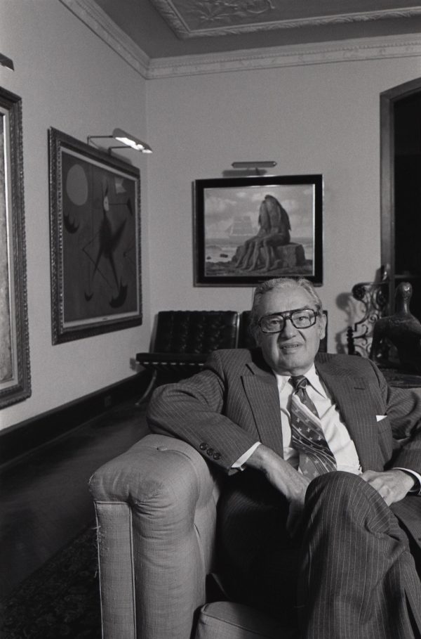 An older gentleman wearing glasses sitting on a chair in a room filled with paintings on the two walls seen behind him