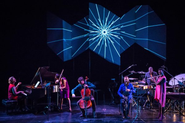 A dynamic concert featuring musicians playing piano, violin, cello, clarinet, percussion, and flute. A striking backdrop displays an abstract blue design resembling a starburst.