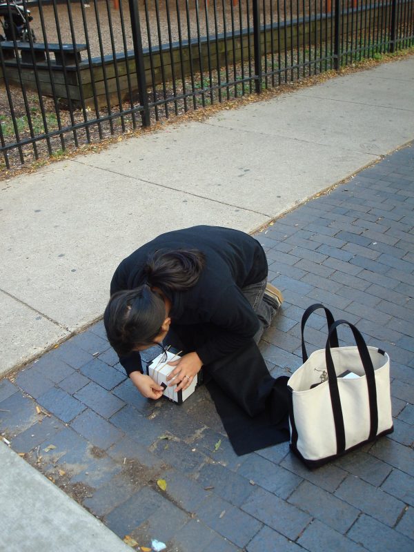 A person dressed in dark clothing kneels, hunched over a small white box and pulling out a small black tab.