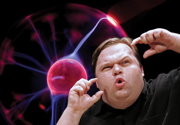 A heavy-set man makes an expressive face with one eyebrow raised and mouth puckered, holding his hands close to his face. Behind him is a clear glass globe emitting pink and purple electricity.