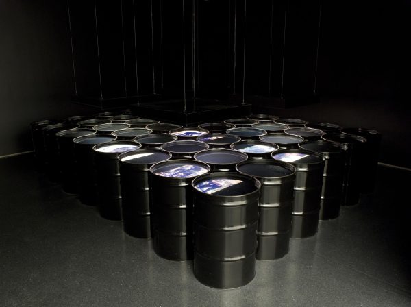 Rows of black barrels in a v formation filled with liquid in a dark room painted black. Images are projected onto the surface of some barrels.