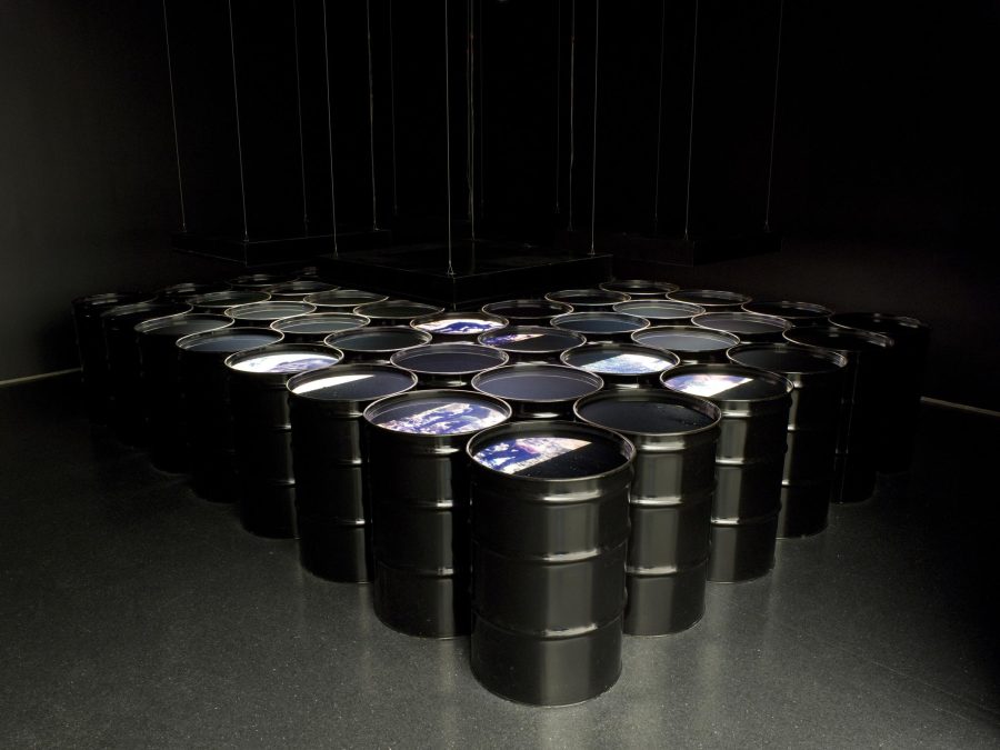 Rows of black barrels in a v formation filled with liquid in a dark room painted black. Images are projected onto the surface of some barrels.