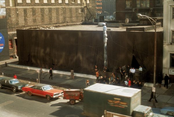 A view of a two-story building that has been wrapped in brown fabric as viewed from higher up. There is a bustle of cars and people around it.