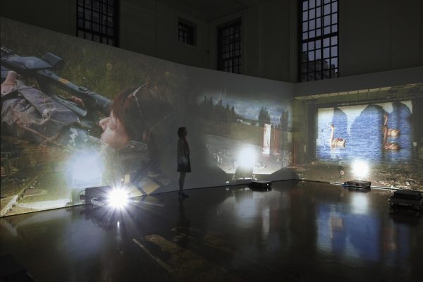 Installation view of a dark room with high ceilings that has three projectors on the floor projecting onto two gallery walls videos of birds swimming on the right and a female hunter aiming a gun on the left. A woman stands in the middle the installation watching the videos.