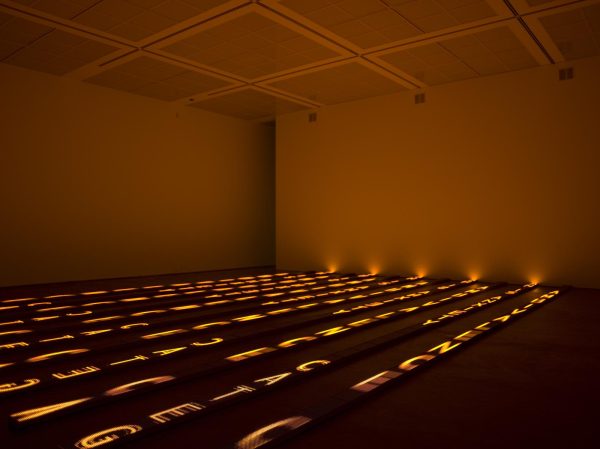 In a large darkened room, large letters are illuminated in yellow in vertical rows on the floor. These letters cast a warm glow over the room.