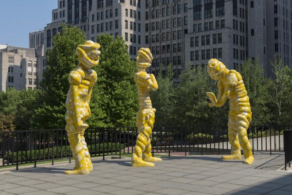 Three yellow sculptures of abstract human figures stand as if communicating with each other outdoors in an urban setting.