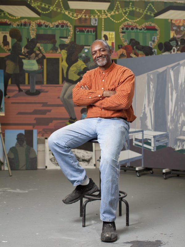 A Black man sits with arms crossed on a tall drafters chair in front of a large painting of people in a salon. His arms are crossed but has a large smile on his face, which sports a salt and pepper beard