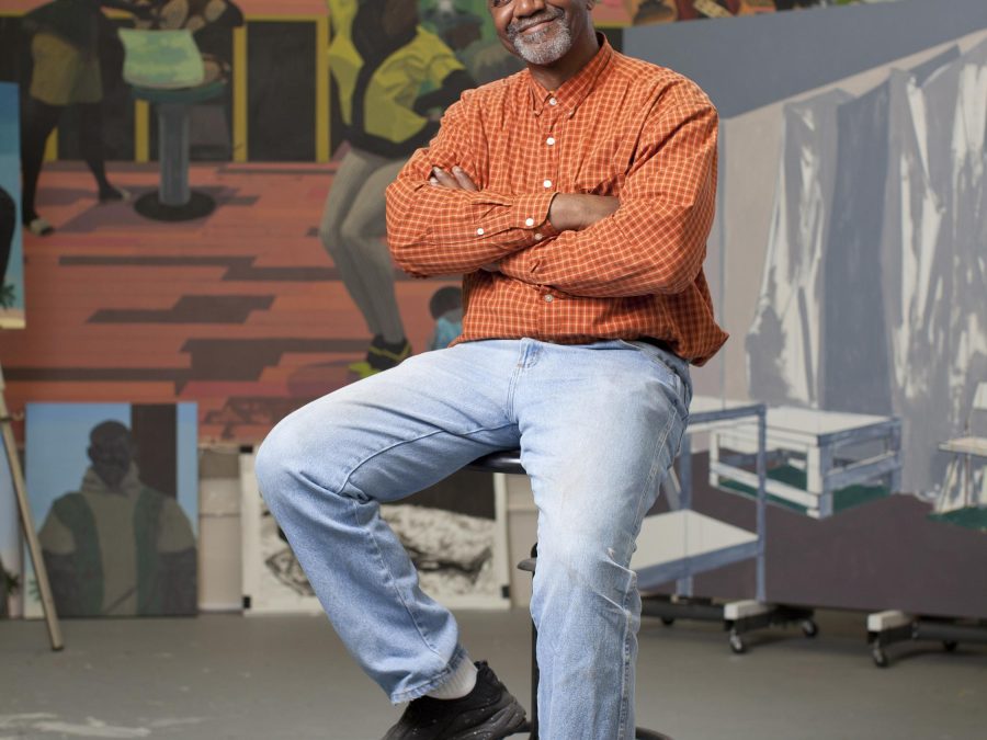 A Black man sits with arms crossed on a tall drafters chair in front of a large painting of people in a salon. His arms are crossed but has a large smile on his face, which sports a salt and pepper beard