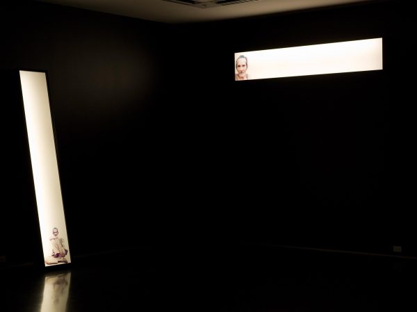 Two narrow glowing rectangles depicting a man illuminate a pitch-black room. The horizontal box on the right hangs high on the wall; the other leans vertically against the wall to the left.