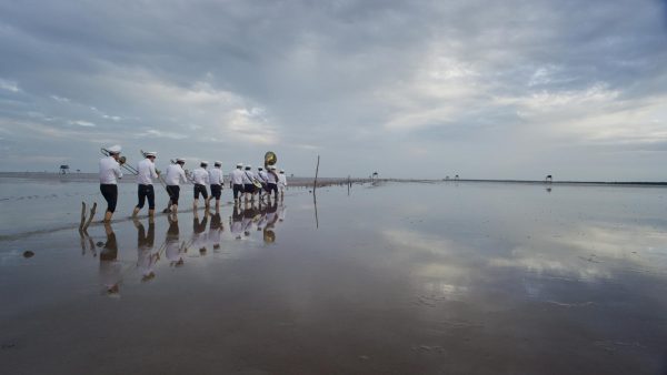 A line of people in uniform hold brass instruments while wading through a body of water