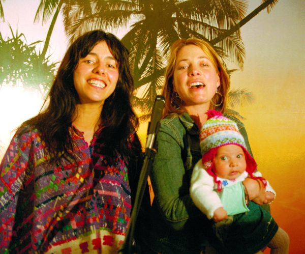 Two female-presenting people, one holding a baby in a winter hat, pose against a tropical backdrop featuring a palm tree
