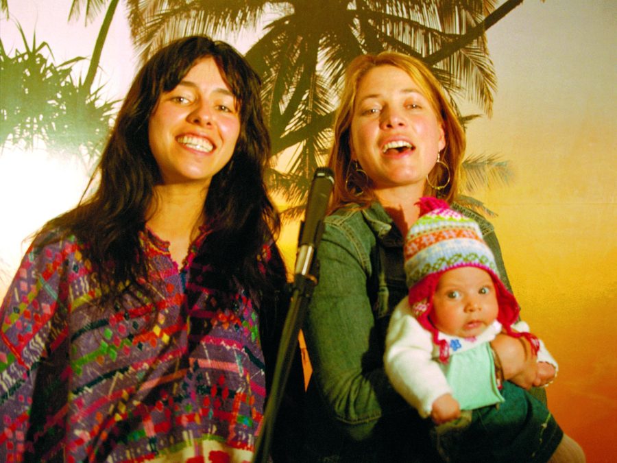 Two female-presenting people, one holding a baby in a winter hat, pose against a tropical backdrop featuring a palm tree