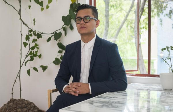 A male presenting person with light-tan skin sits at a marble table in a suit with a trailing plant that has been tacked to the white wall behind him