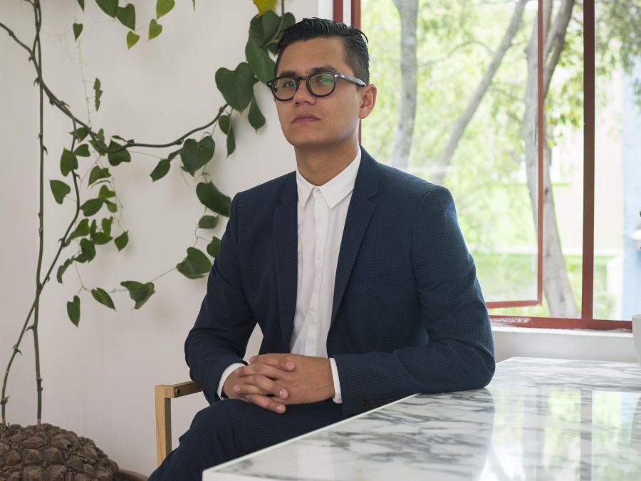 A male presenting person with light-tan skin sits at a marble table in a suit with a trailing plant that has been tacked to the white wall behind him