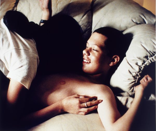 A shadowed figure leans over a partially nude youth, who is smiling with his eyes closed. They are both in a bed with gray sheets.