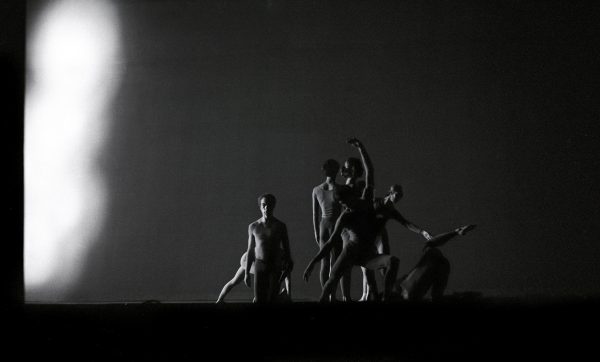 The black-and-white image captures a cluster of seven dancers holding poses against an empty background, with a strong light coming from the left.