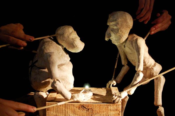 Two clay puppets with sticks attached to their heads and hands interact at a wooden block