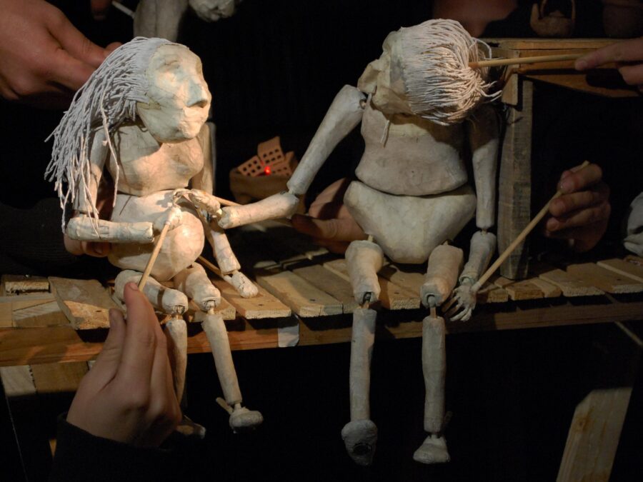 Two humanoid puppets hold hands while seated on a tiny wooden stage.