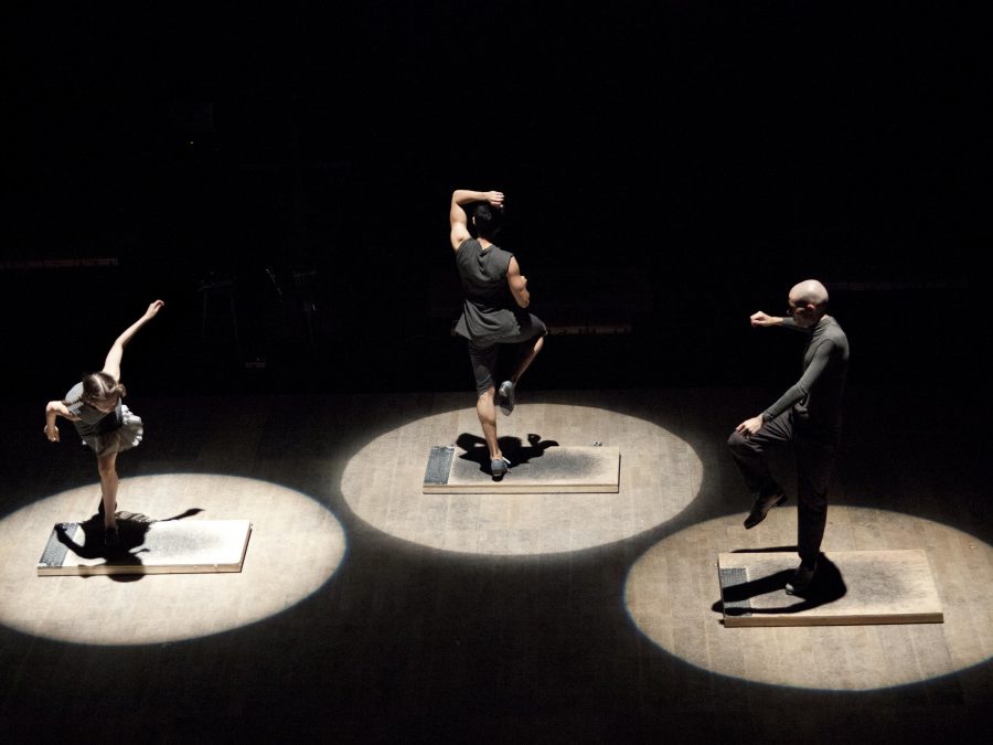 Three tap dancers dressed in black and gray—illuminated by spotlights—dance atop individually sized platforms. Each faces a different direction, and seems to be in he or her own dance.