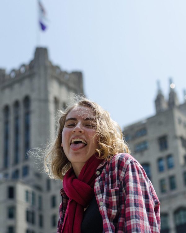 A light-skinned person with wavy blond hair sticks their tongue out while looking down towards the camera outside of a tall collegiate-looking building