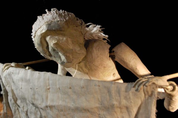 A roughly made puppet with long white fabric hair tilts their head toward a piece of fabric that resembles a sheet hanging on a laundry line. Their hands rest on either side of the fabric.