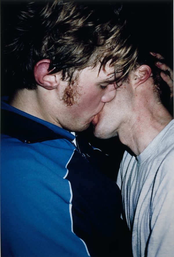 Close up of two pale-skinned, male-presenting people sharing a kiss.