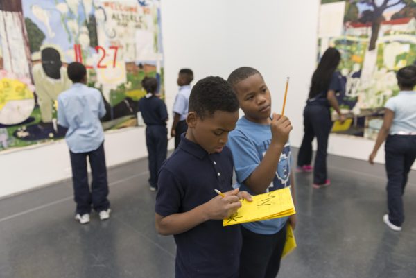 Students in a gallery space looking at large paintings. In the foreground two students engage with each other as one writes in a yellow journal