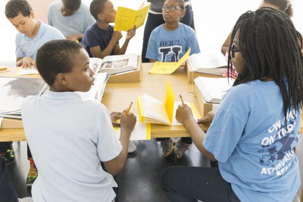Kids gather at a table holding or writing in yellow journals