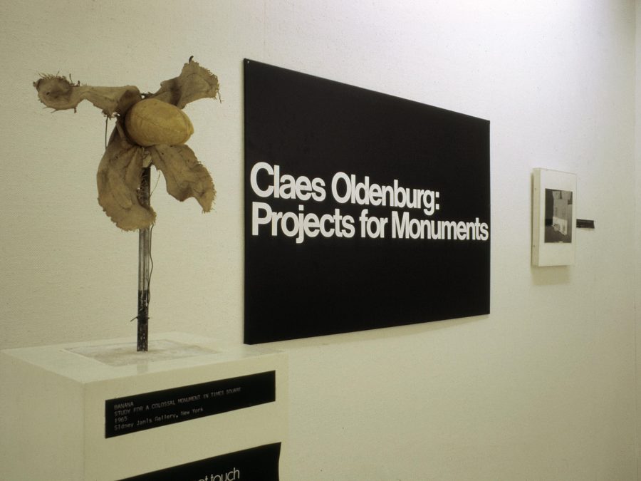 A small sculpture held up by metal on a pedestal next to a black rectangle with white text reading Claes Oldenburg Projects for Monuments