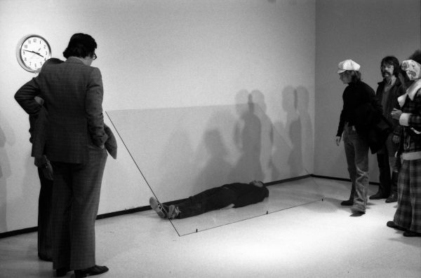 A group of people observe a man lying on the floor underneath a sheet of glass.
