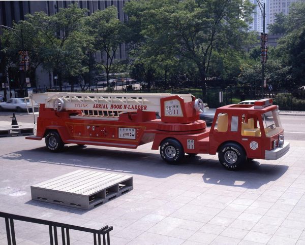 A red firetruck parked on a plaza across from a park with giant gray pallets in front of it
