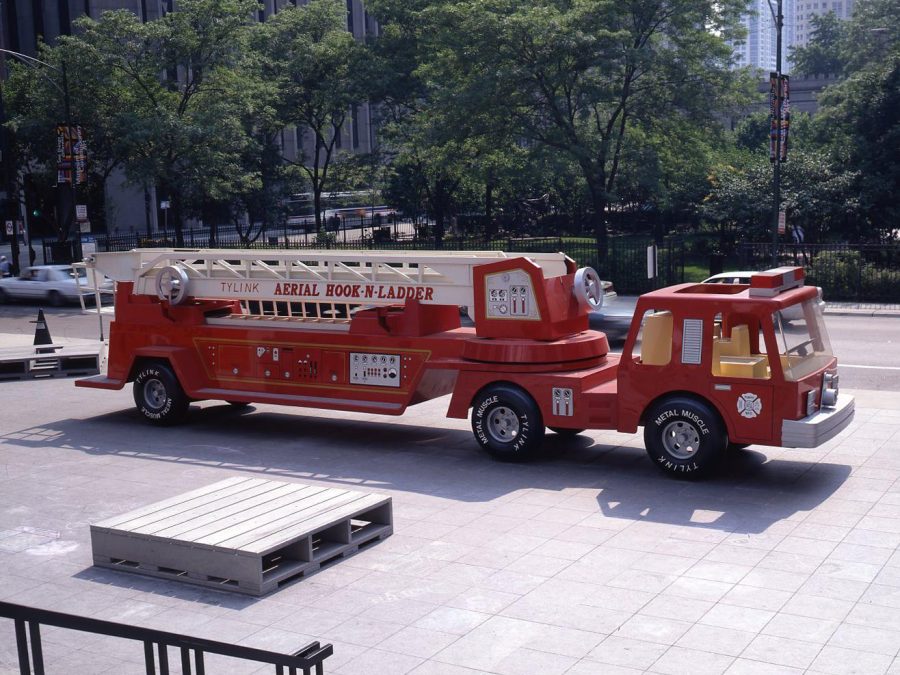 A red firetruck parked on a plaza across from a park with giant gray pallets in front of it