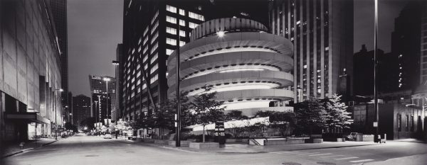 Black and white view down a city street at night featuring a circular ramp next to a building with all of its lights on on the right side