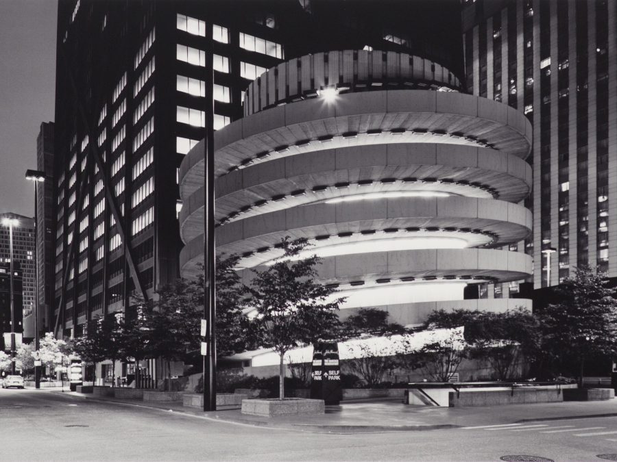 Black and white view down a city street at night featuring a circular ramp next to a building with all of its lights on on the right side