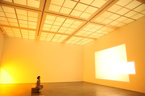 A person looks at a bright projection from a bench in an otherwise empty room lit soft orange-yellow