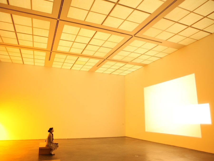 A person looks at a bright projection from a bench in an otherwise empty room lit soft orange-yellow