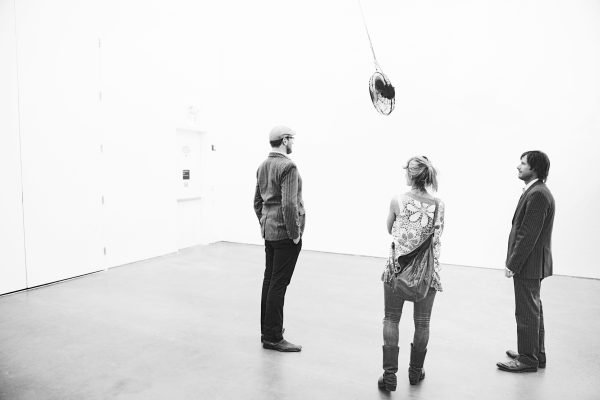 Three people in a black-and-white photograph look at a ceiling fan hanging from the ceiling.