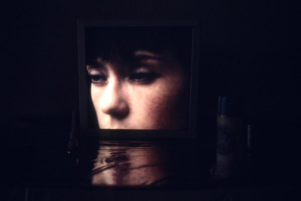 A projection of a person's head, cut off at the mouth down, reflects onto the floor of a dark room