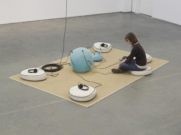 A person sits on a white cushion on a carpet in a large stark room with three other cushions at each corner and headphones resting on top of each. Cords lead to the headphones from a blue ball in the middle.